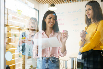 Smiling female professionals writing on glass wall at office
