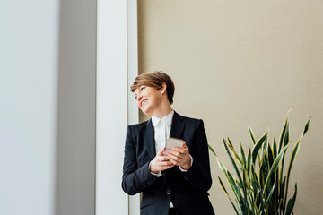 Happy female business professional looking away while holding mobile phone against beige wall in office