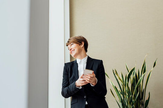 Happy female business professional looking away while holding mobile phone against beige wall in office