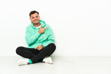 Young handsome caucasian man sitting on the floor pointing finger to the side