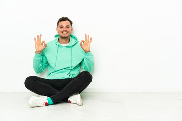 Fototapeta premium Young handsome caucasian man sitting on the floor showing an ok sign with fingers