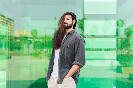 Young male hipster with long brown hair standing in front of glass wall