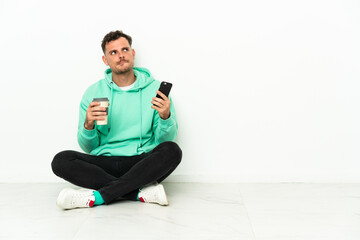 Obraz premium Young handsome caucasian man sitting on the floor holding coffee to take away and a mobile while thinking something