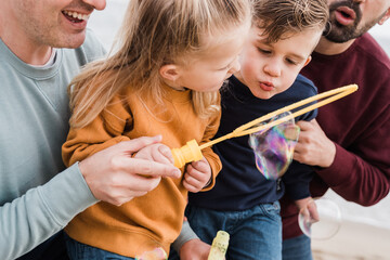 Gay male couple and kids having fun on the beach during summer vacation using soap bubble toy - Lgbt and love family concept - Main focus on right brother face