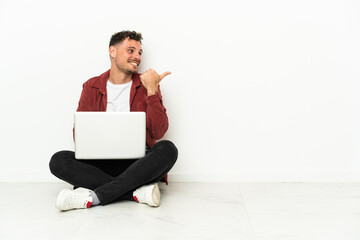 Young handsome caucasian man sit-in on the floor with laptop pointing to the side to present a product