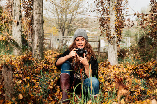 Smiling woman photographing through camera while sitting amidst autumn leaves by dog