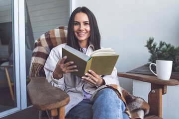 Smiling woman holding book while sitting on chair on balcony