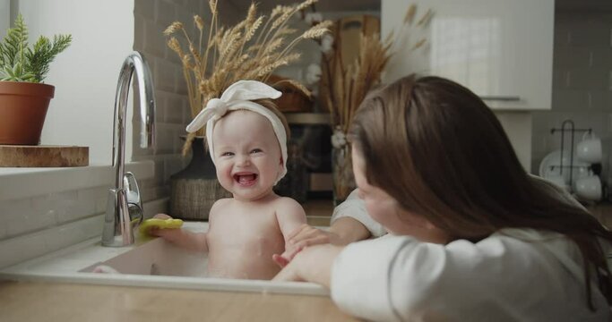 Mother and daughter spend time at home together. Young mom helps her adorable little child to take a bath in a kitchen sink. Adorable girl have fun in the water and foam.