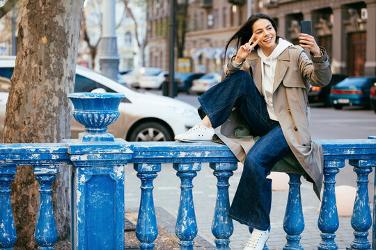 Smiling woman sitting on blue railing showing peace sign while taking selfie