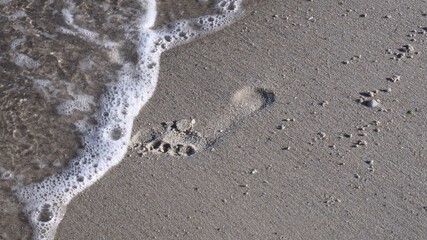 Sea surf at clean white sand with footprint of barefoot legs. Waves erase footprint from beach sand. Coastal vacation background with copy space