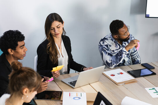 Businesswoman with laptop reading adhesive note while sitting by colleagues in meeting