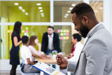 Young businessman checking document on clipboard with colleagues in background at workplace