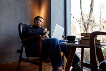 A young girl with glasses, with short hair is sitting in a cafe and working on a laptop. The girl looks at her laptop and makes notes in a notebook. The concept of distance learning