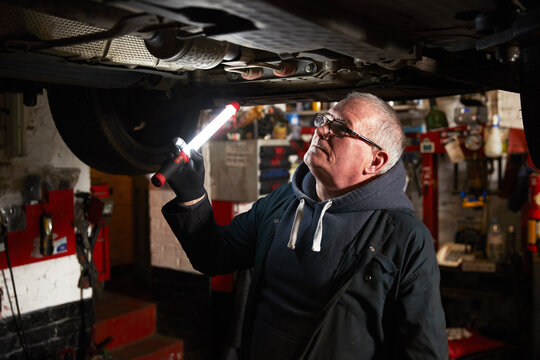 Senior male mechanic with flashlight examining car in repair shop