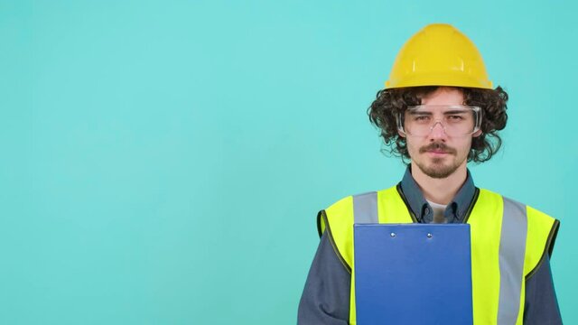 Handsome young construction worker with safety glasses and helmet that is thinking while holding clipboard and looking at camera against turquoise background, left copy space.