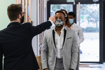 Man wearing face mask checking temperature of colleague with infrared thermometer at office