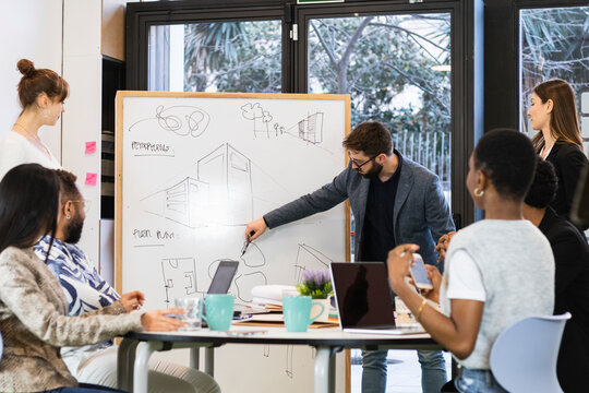 Young entrepreneur explaining over diagram on whiteboard to colleague in meeting