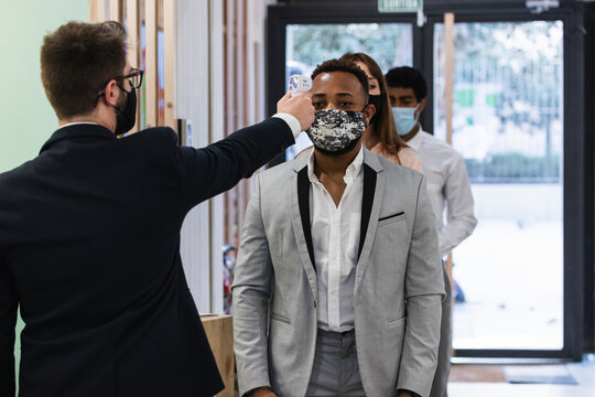 Man wearing face mask checking temperature of colleague with infrared thermometer at office