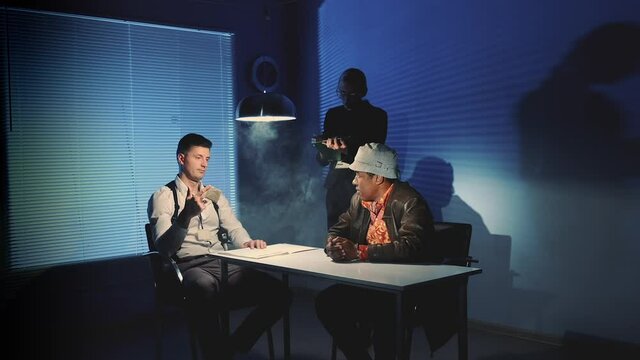 Interrogation room in smoke: detective showing evidence of drug trafficking to suspected black man. Handcuffed man denying charges.