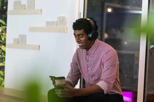 Smiling male professional with headphones using smart phone while having coffee in office