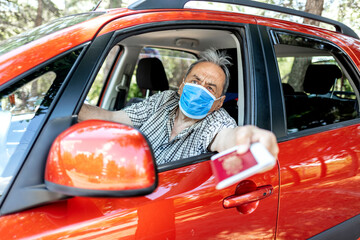 Senior with protective face mask sitting in car passing his identification documents. Man...
