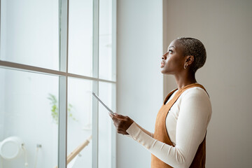 Contemplating woman holding digital tablet while standing in front of window at home