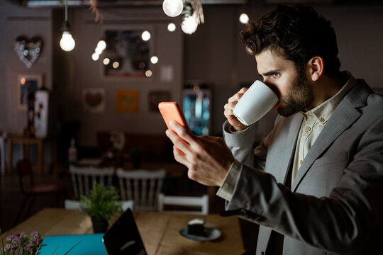 Mid adult businessman working on mobile phone while drinking coffee in cafe