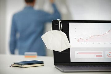 Face mask hanging on laptop screen while man standing in background at office