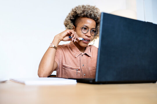 Female entrepreneur biting pen while working on laptop at home office