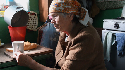 Side view portrait of 80 years old woman having breakfast in rustic kitchen. Rural senior woman in traditional head scarf wrapped on her head. Life after retirement