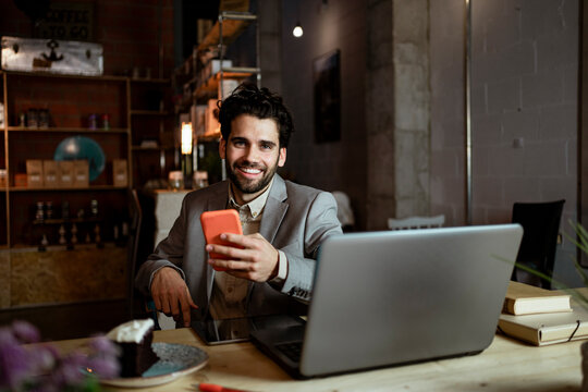 Happy businessman with laptop holding mobile phone while sitting in coffee shop