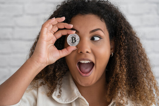 Excited woman with mouth open covering eye with bitcoin in front of wall