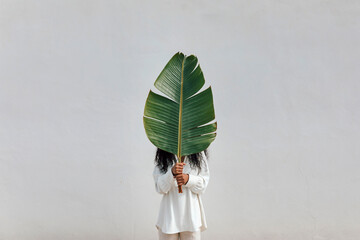 Woman covering face with big green leaf in front of white wall