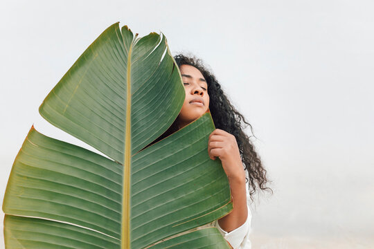 Curly haired woman with eyes closed holding big green leaf in front of white wall