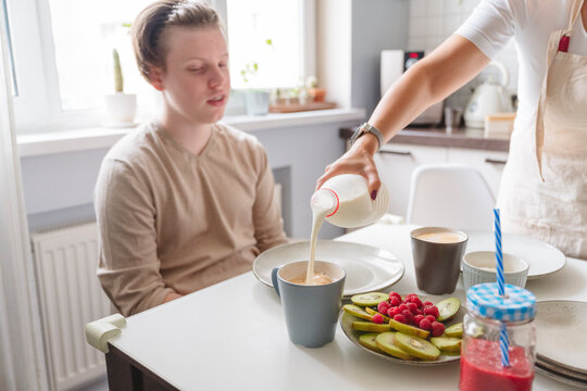 Woman pouring milk in cup for teenage son at dining table in kitchen