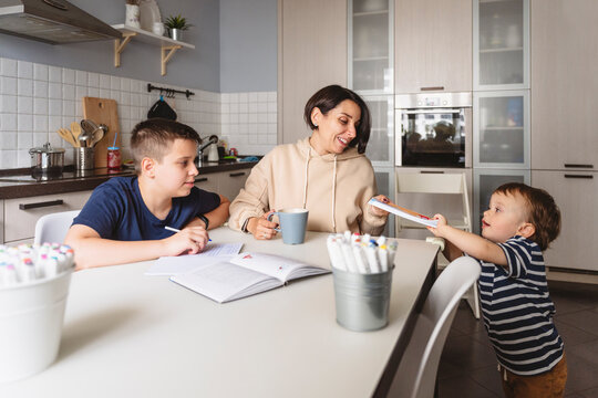 Boy giving book to mother while brother doing homework at table in kitchen