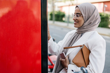 Smiling woman with purse holding smart phone while using self service machine by road