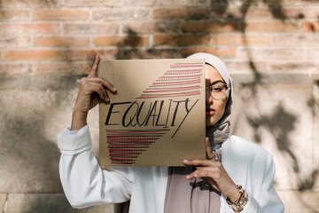 Young woman covering face with placard in front of wall on sunny day