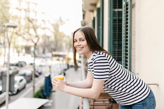 Happy female tourist with glass of juice leaning on railing in balcony