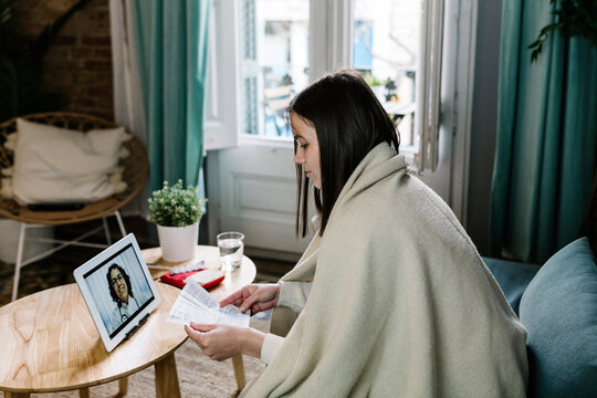Sick woman wrapped in blanket talking during telemedicine through digital tablet at home