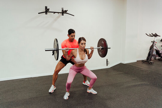 Fitness instructor guiding female athlete while exercising with barbel in gym