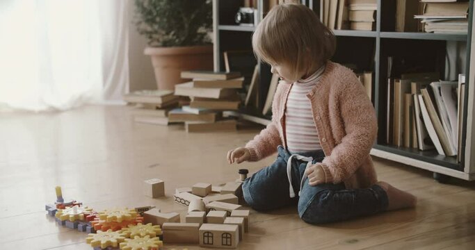 Cute toddler girl sits near wooden blocks, firstly she looks, secondly she starts to play. Toy playful building. Assembling a tower of wooden bars. Close up wooden puzzle blocks, light window