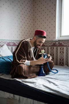 Smiling man with hookah using smart phone on bed at home