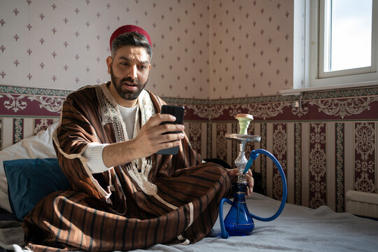 Bearded man using mobile phone while sitting with hookah on bed at home