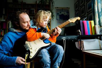 Father teaching electric guitar to blond son while sitting at home