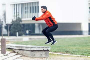 Sporty man doing jumping squats on retaining wall at public park