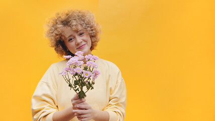 Beautiful girl with curly hair holding a bunch of purple flowers. Girl smiling and in a good mood dressed in a baggy shirt. Isolated over bright yellow background studio. High-quality photo 