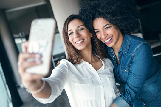 Cheerful businesswoman embracing female colleague while taking selfie through mobile phone in office