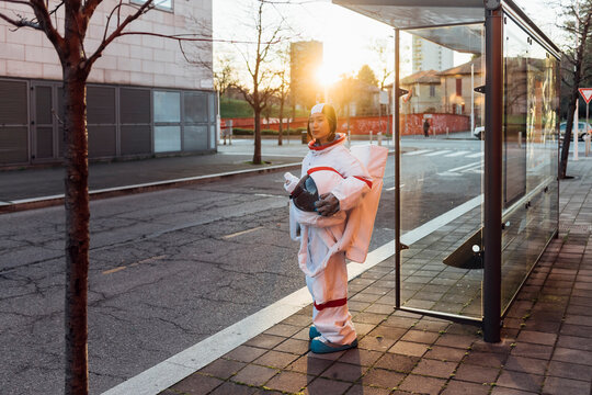 Female astronaut with space helmet standing at bus stop during sunset
