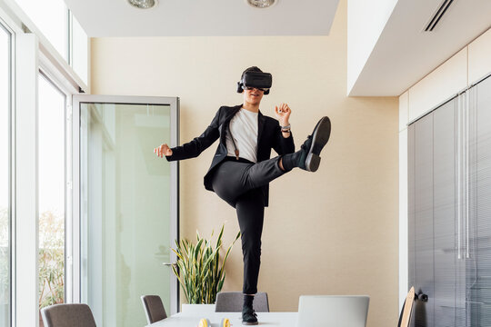 Young female entrepreneur with virtual reality headset kicking while standing on desk in office